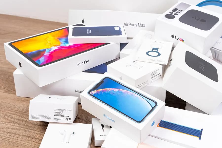一堆苹果产品的包装盒——包括iPhone、iPad Pro、AirPods、Apple Watch和Apple TV——整齐地摆放在木质台面上。