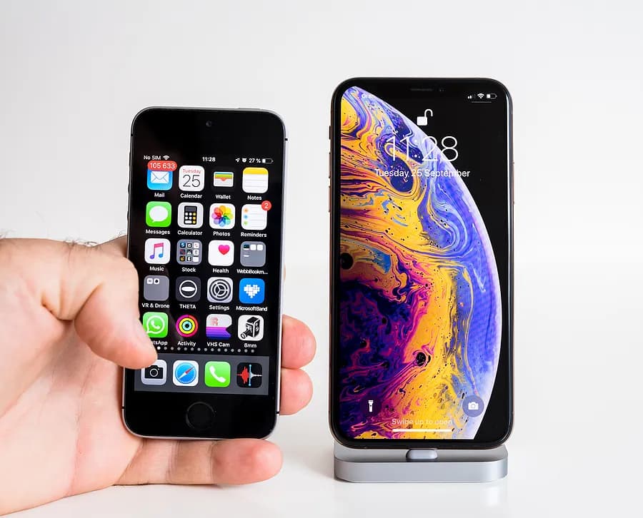 一只手拿着一部较旧的小型iPhone,将其与支架上的一部较大的现代iPhone并排放置,以此展示旧款iPhone与新款iPhone在尺寸和设计上的差异。